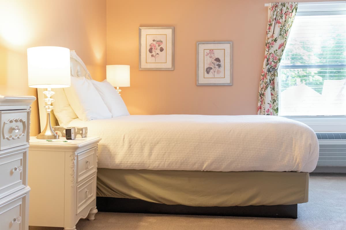 Een net opgemaakt bed met een witte deken en een donkere ondergrond, een witte nachtkastje met een lamp, en twee lijstjes met bloemen op de muur.