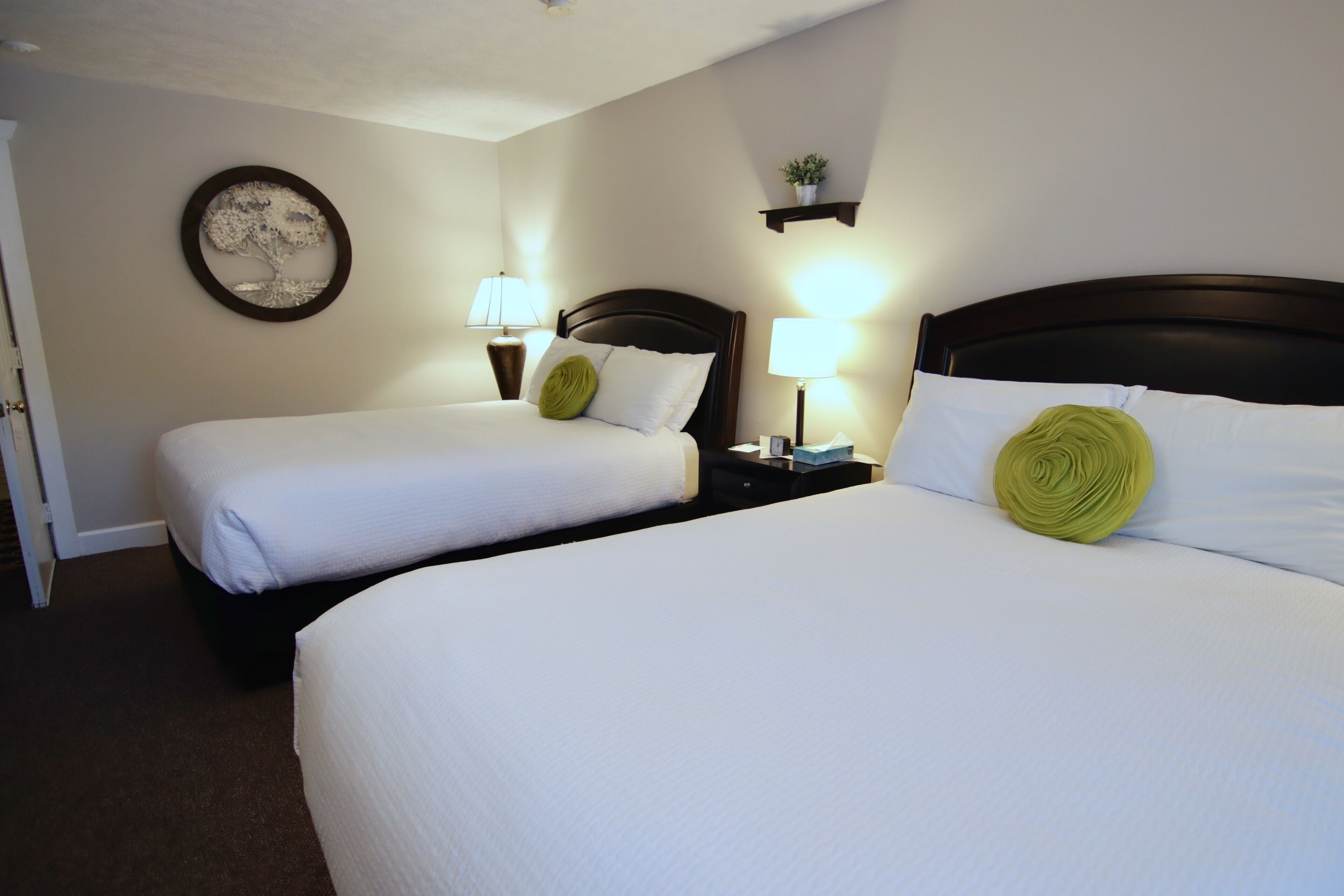 A hotel room with two beds, each with white bedding and green decorative pillows. There are bedside lamps and a wall-mounted shelf with a plant.