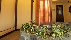 A stone pillar with a copper base surrounded by greenery, supported by a wooden ceiling with hanging lights.