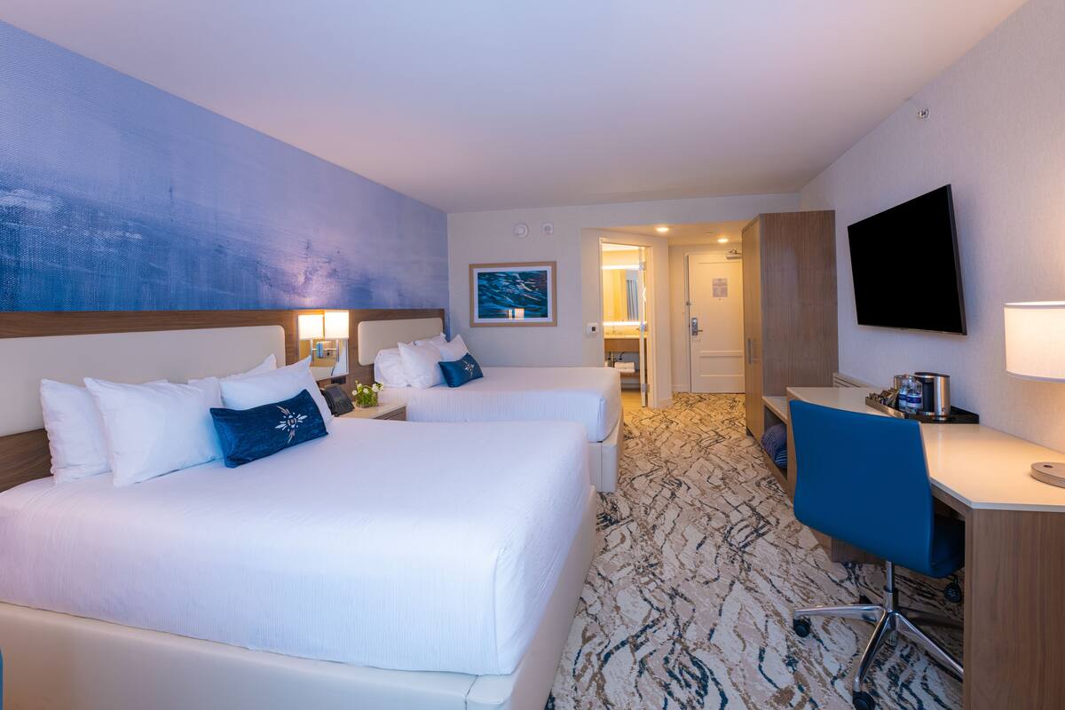 A hotel room with two beds, a desk, a TV, and a bathroom visible through an open door.