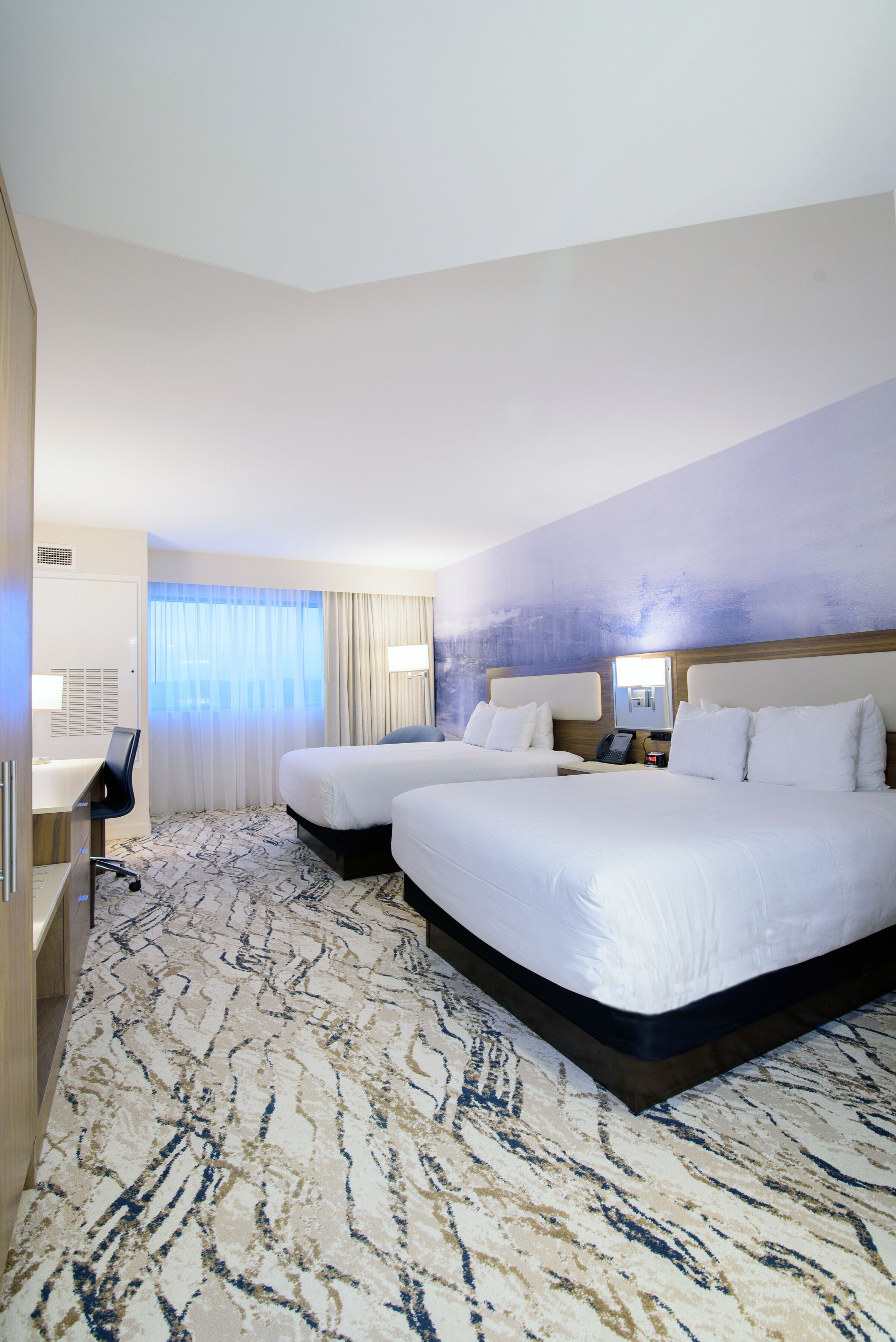 A hotel room with two beds, a desk, and a large window with curtains.