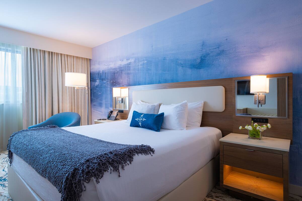 A hotel room with a large bed, a bedside table with a lamp, a chair, and a window with curtains.