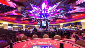 A casino floor with multiple blackjack tables, a bar area, and a vibrant ceiling with geometric light patterns.