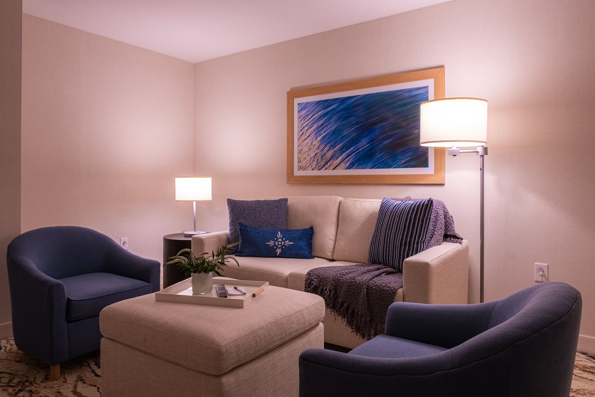 A living room with a beige sofa, two blue armchairs, a coffee table with a tray, a lamp, and a painting on the wall.