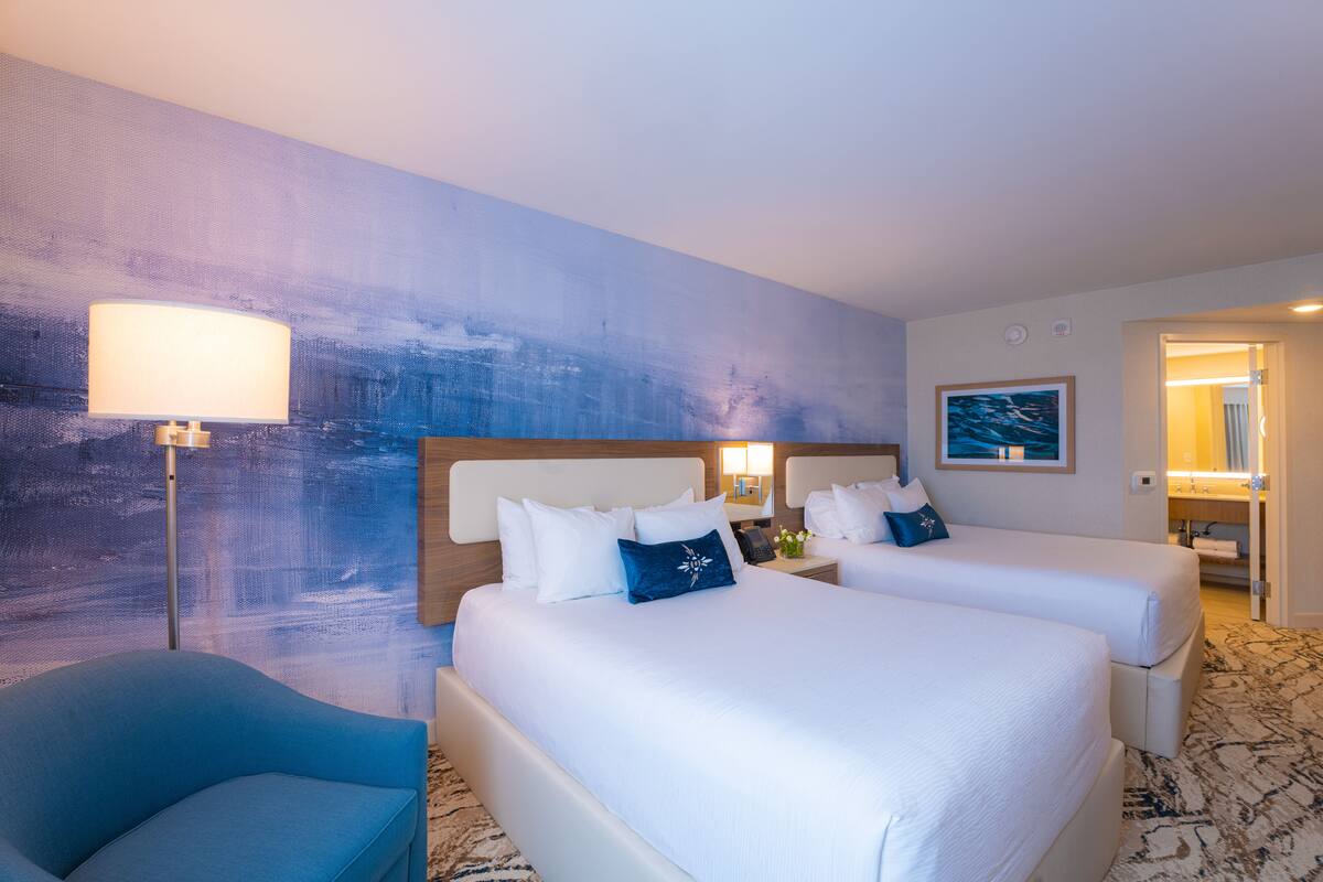 A hotel room with two beds, a blue armchair, a large abstract painting on the wall, and a bathroom visible through an open door.