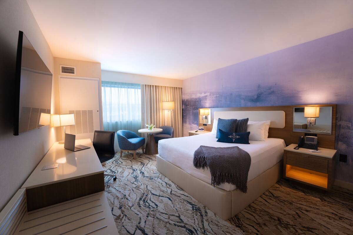 A hotel room with a large bed, bedside tables, a desk, a chair, and a small round table.