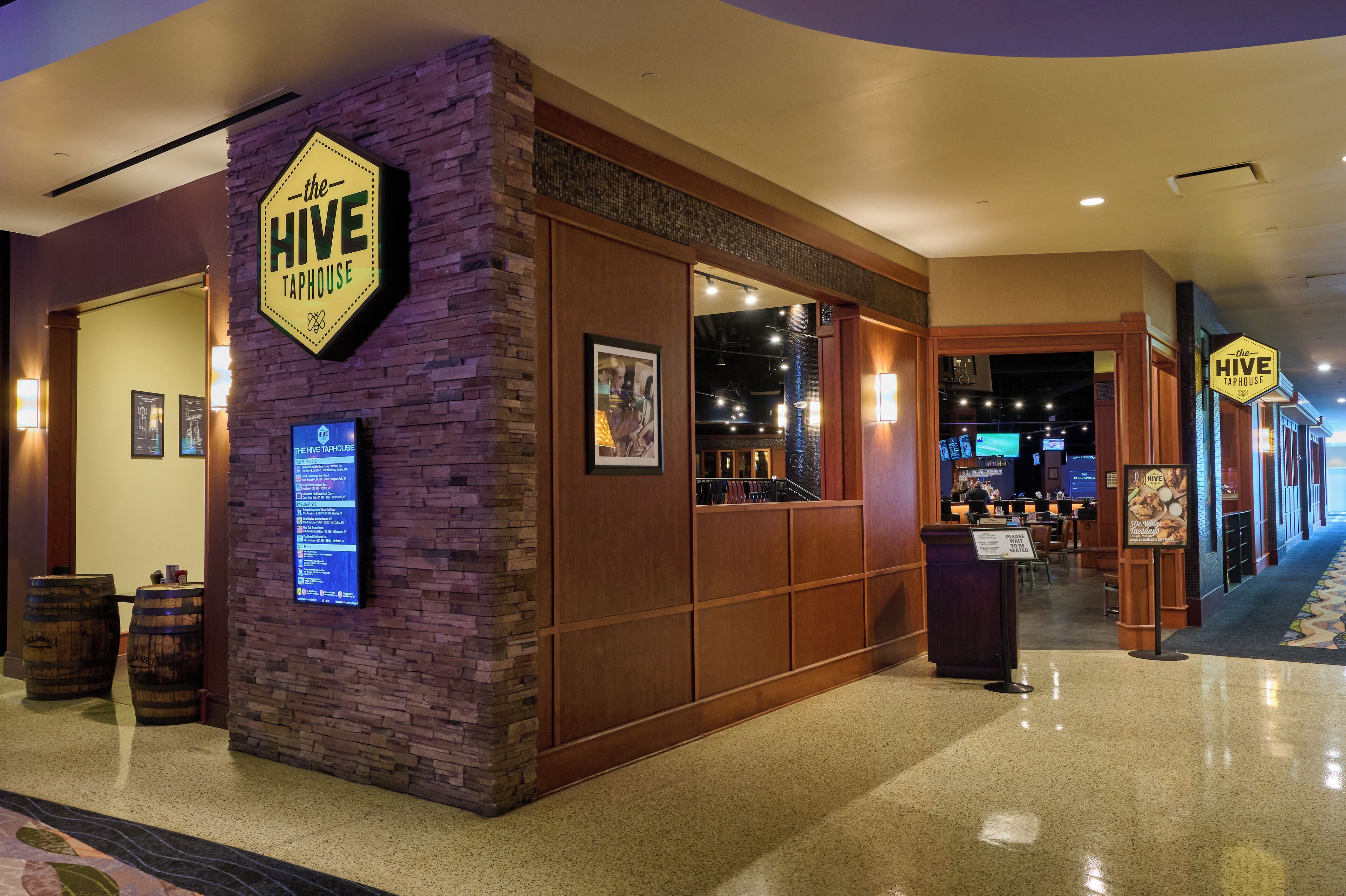 Un hall d’hôtel avec une enseigne indiquant « The Hive Taphouse », des panneaux de bois, un menu numérique et un bar visible à travers la vitre.