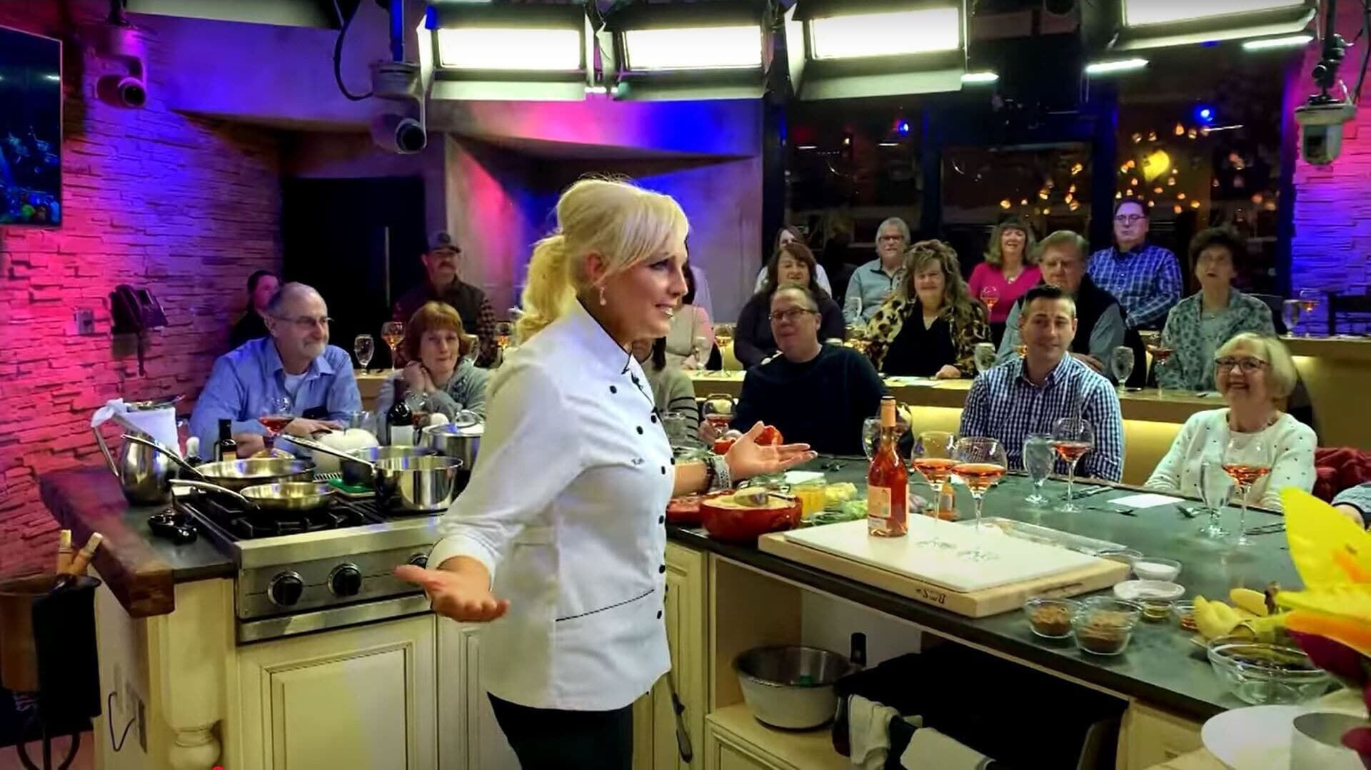 Un chef, vêtu d’une blouse blanche, cuisine dans une cuisine, tandis qu’un groupe de personnes l’observe.