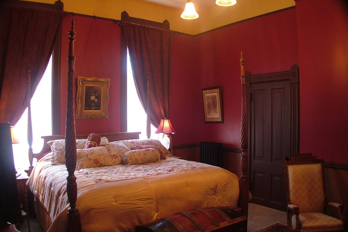 A four-poster bed with a canopy, a chair, and a door in a room with red walls and a wooden floor.