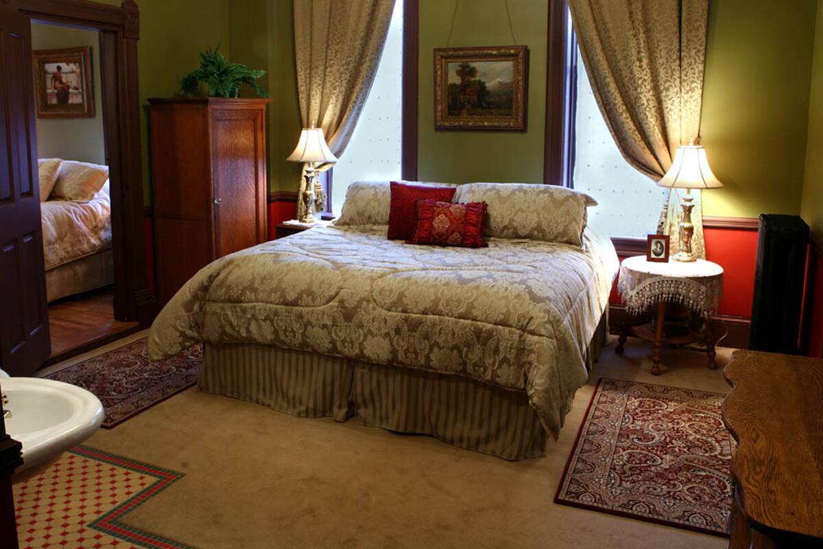 A bedroom with a large bed, two bedside lamps, a nightstand, a small table, a mirror, and a door leading to another room.