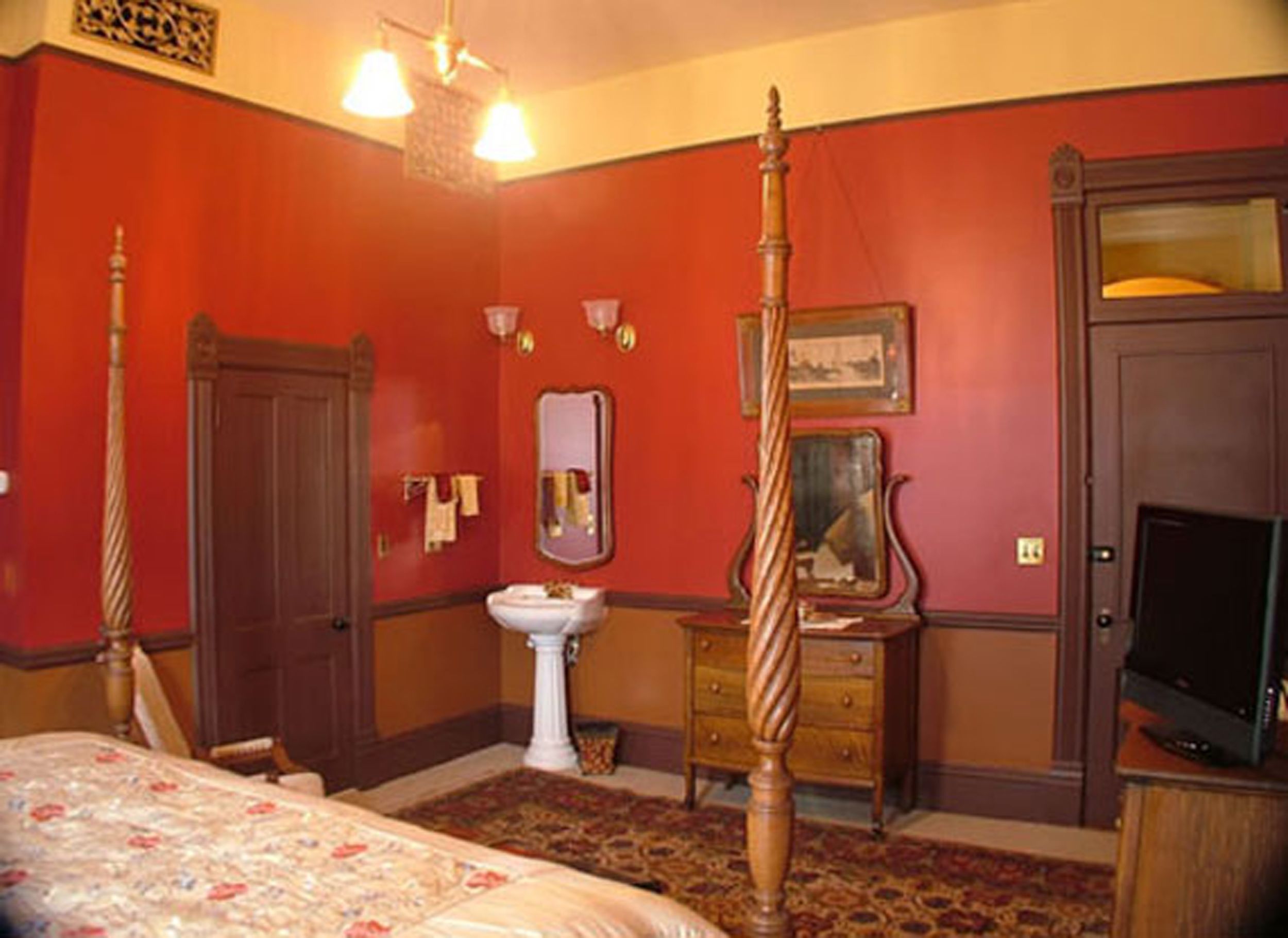 A bedroom with a four-poster bed, a wooden wardrobe, a bedside table with a mirror, a white pedestal sink, and a television on a wooden stand.