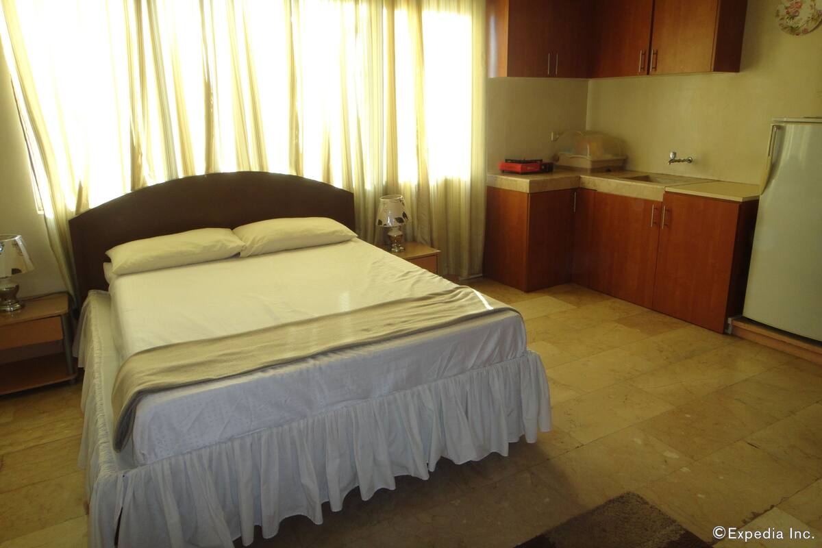 A hotel room with a bed, a nightstand, a small table, a kitchen area, and a refrigerator.