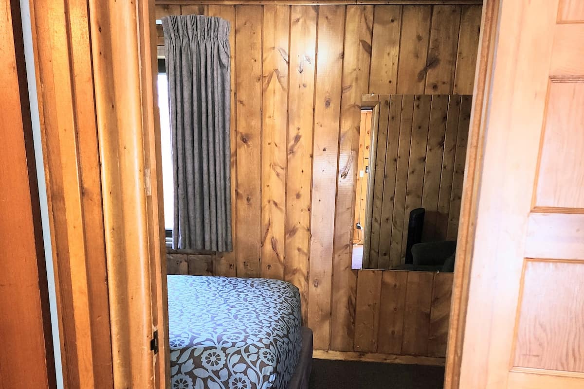 Ein Zimmer mit Holzwänden, einem Bett mit gemusterten Textilien, einem Fenster mit Vorhängen und einem Spiegel an der Wand.