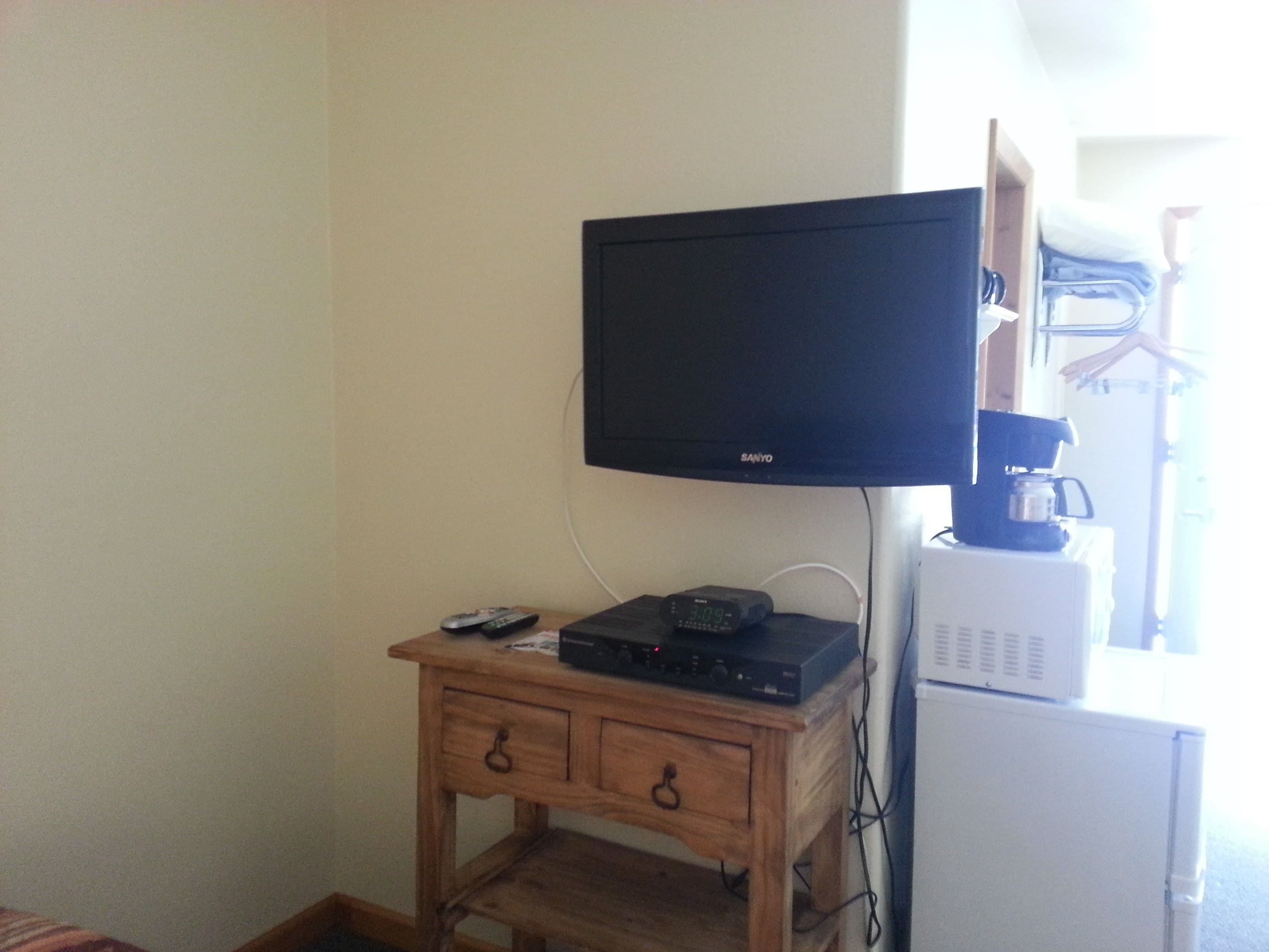 A room with a wooden desk, a black television mounted on the wall, a coffee maker, and a white refrigerator.