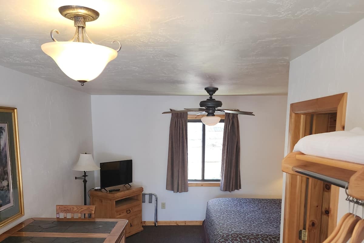 Ein kleines Zimmer mit Bett, Schreibtisch mit Stuhl, Fernseher, Deckenventilator und einem Fenster mit Vorhängen.