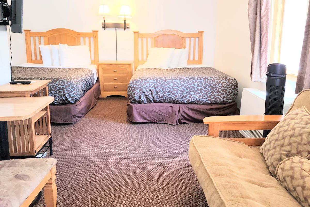 Ein Zimmer mit zwei Betten, einem Schreibtisch, einem Stuhl und einer Couch. Über den Betten befinden sich Wandlampen und an der Wand ist ein Fernseher angebracht.