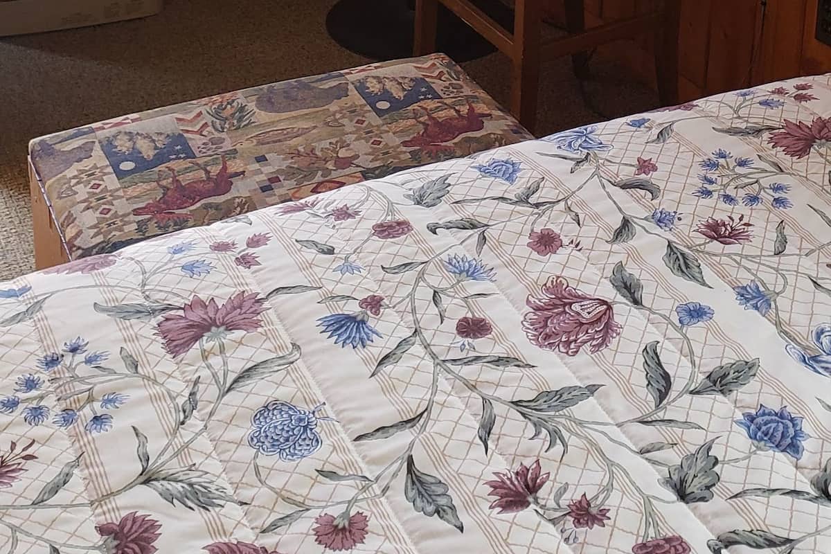 Ein Zimmer mit Holzwänden, einem Wand-Flachbildfernseher, einem Tisch mit einer Speisekarte, einem Korbstuhl, einem Bett mit floralem Überwurf und einem Heizkörper auf dem Boden.