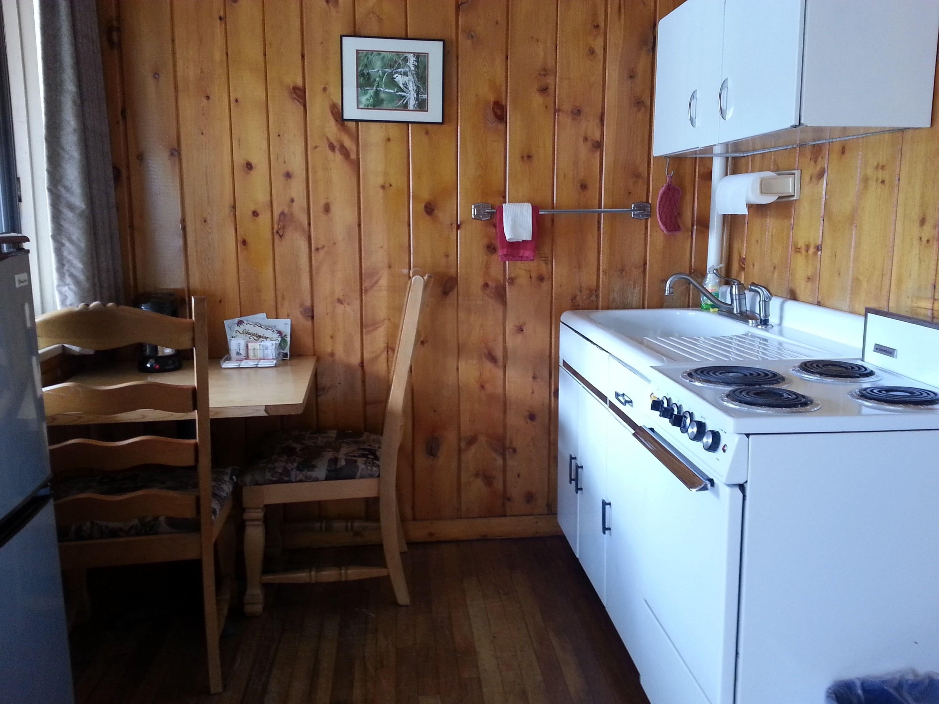 Cocina compacta con paredes de madera, estufa blanca, una silla de madera y una pequeña mesa con algunos objetos encima.