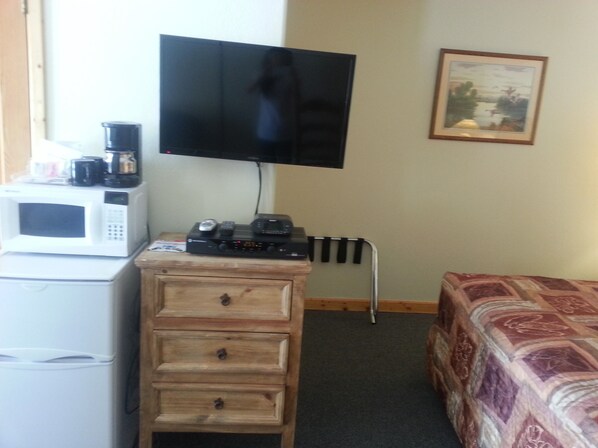 A room with a TV mounted on the wall, a microwave on a cabinet, a small table with a coffee maker, and a bed with a patterned bedspread.