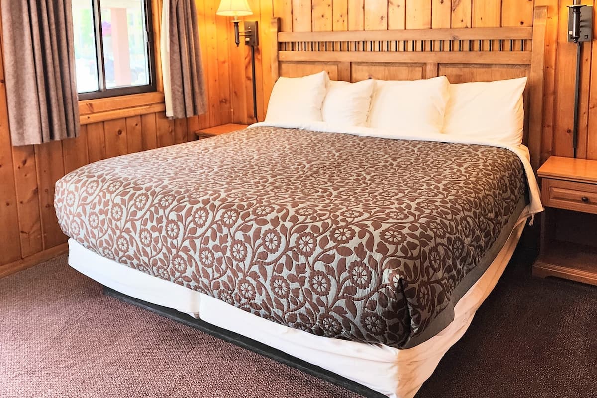 Ein Zimmer mit Holzwänden, einem großen Bett, zwei Nachttischlampen und einem Fenster mit Vorhängen.