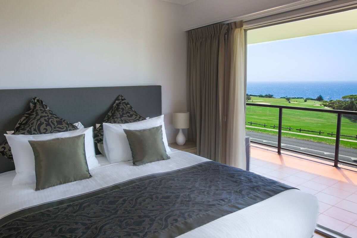 Ein Hotelzimmer mit einem großen Bett, zwei Kissen, einer Nachttischlampe und Blick auf einen Golfplatz und den Ozean.