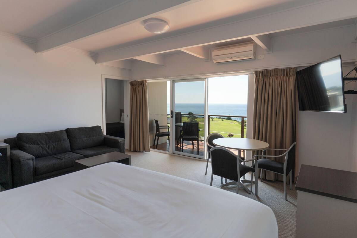 Ein Hotelzimmer mit einem großen Bett, einer Couch, einem Esstisch mit Stühlen, einem an der Wand befestigten Fernseher und einer Glasschiebetür, die auf einen Balkon mit Meerblick führt.