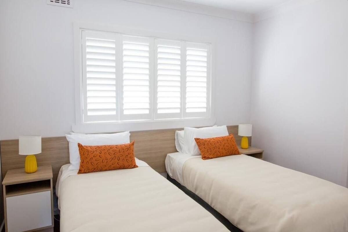 Ein Hotelzimmer mit zwei Betten, weißer Bettwäsche, orangefarbenen Kissen, einem Fenster mit Jalousien und zwei Nachttischlampen.
