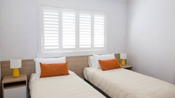A hotel room with two beds, white bedding, orange pillows, a window with blinds, and two bedside lamps.