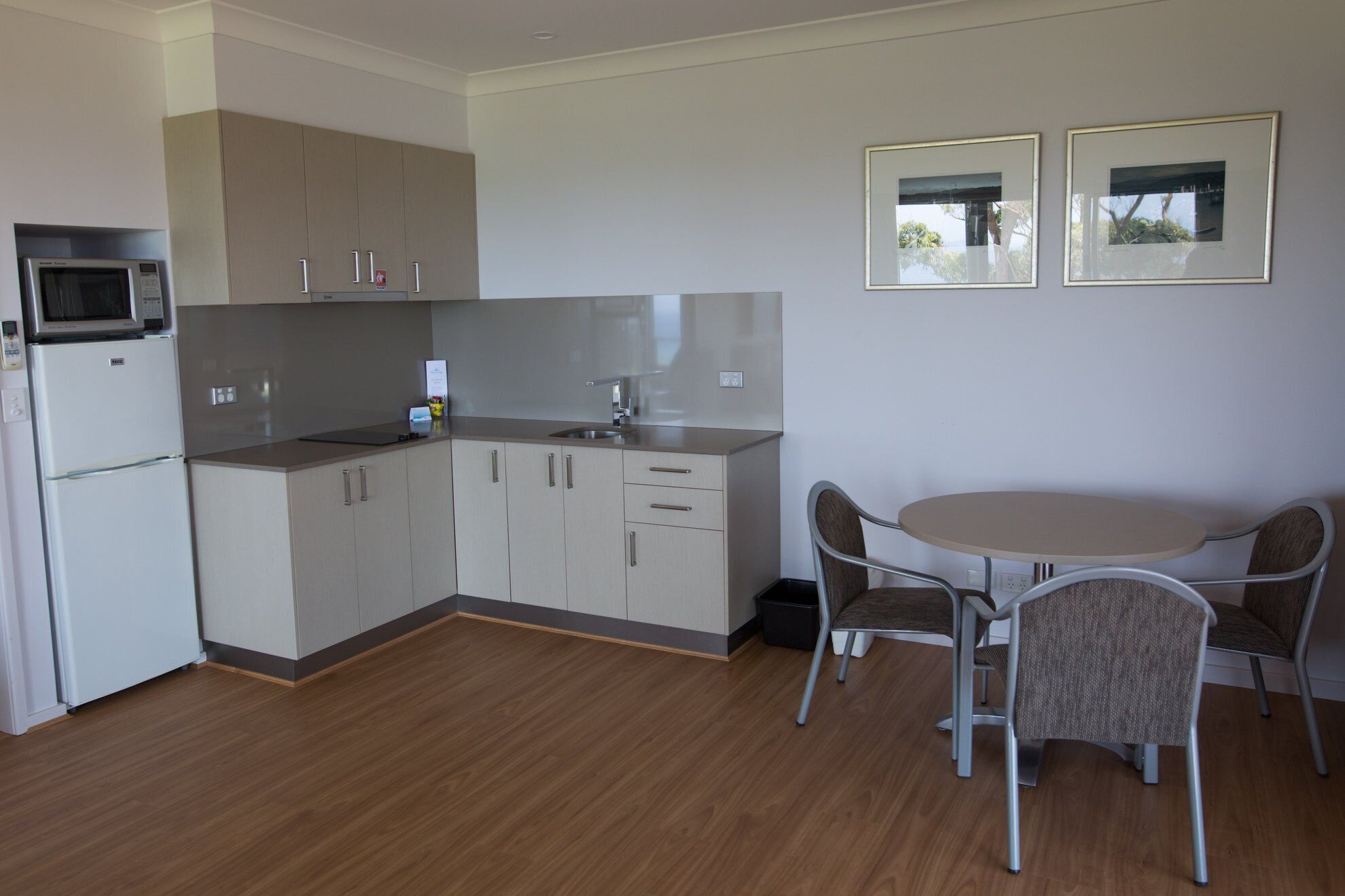 A compact kitchen with a microwave, refrigerator, and a sink. There is a dining table with four chairs and two framed pictures on the wall.