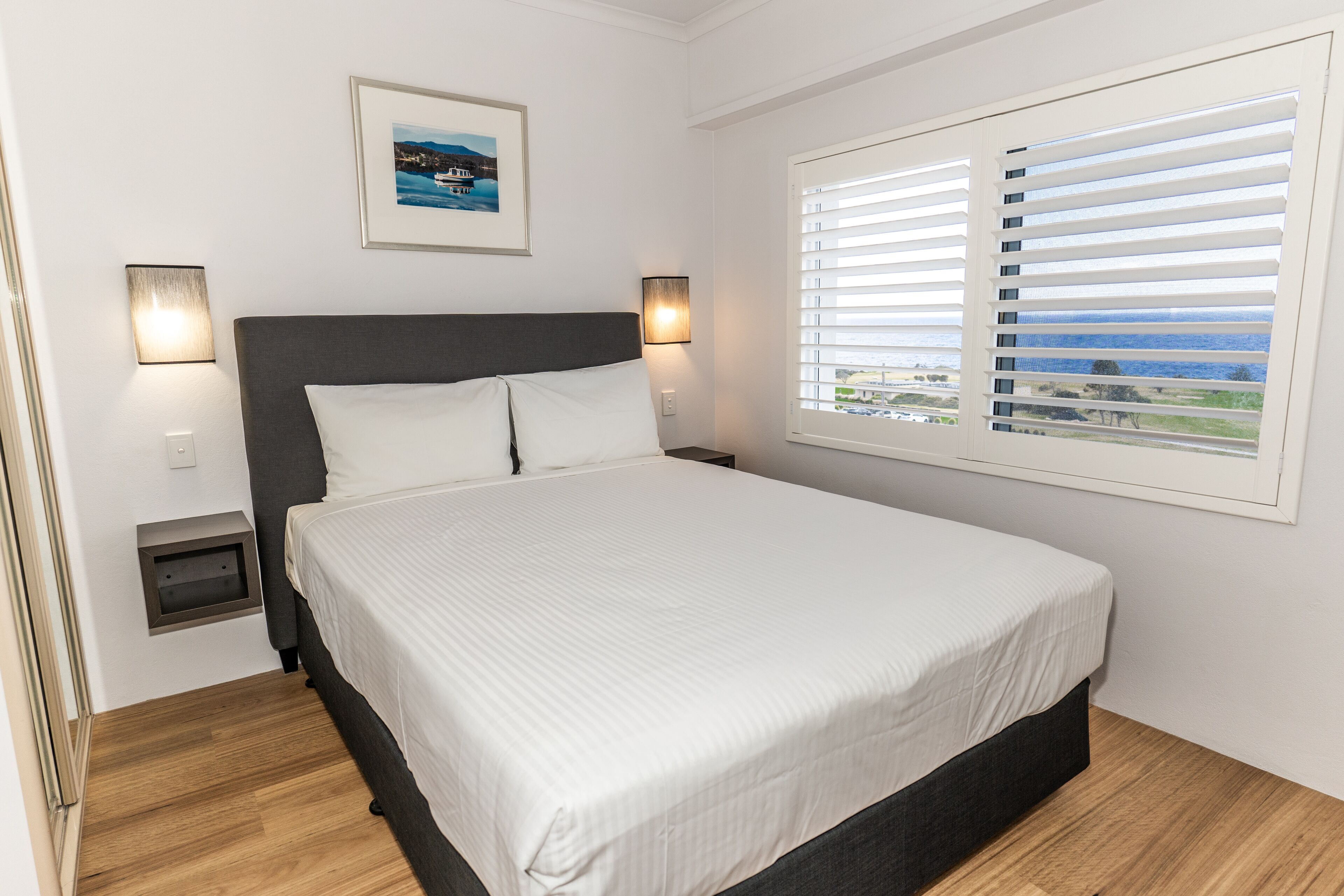 Quarto com cama grande, duas luminárias de cabeceira, quadro emoldurado na parede e janela com persianas.
