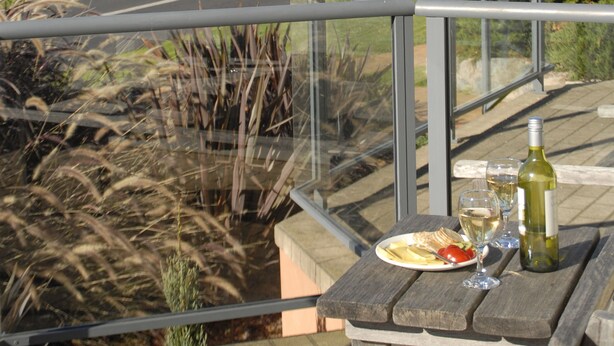 A wooden table with a bottle of wine, a glass, and a plate of food on a balcony overlooking a coastal landscape with a clear sky. - Amooran Oceanside (Narooma)