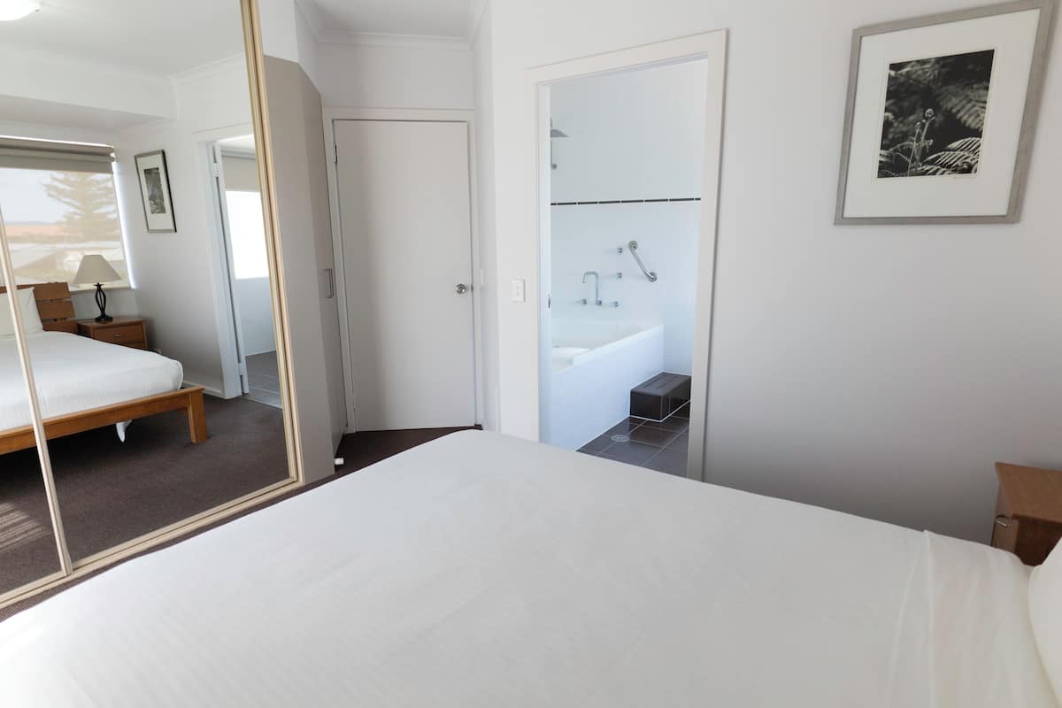 Ein modernes Hotelzimmer mit einem großen Bett, einem Nachttisch mit Lampe, einem Badezimmer mit Badewanne und einem gerahmten Bild an der Wand.