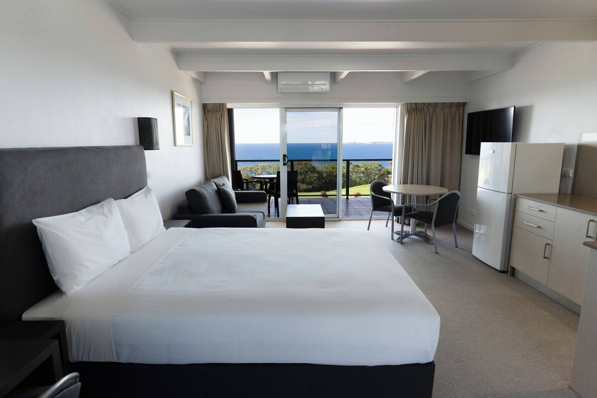 Ein Hotelzimmer mit einem großen Bett, einem Schreibtisch, einem Sessel, einem kleinen Tisch, einem Fernseher und einem Balkon mit Meerblick.
