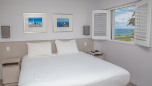 A bedroom with a bed, two framed pictures, a nightstand, a lamp, and a window with shutters. - Amooran Oceanside (Narooma)