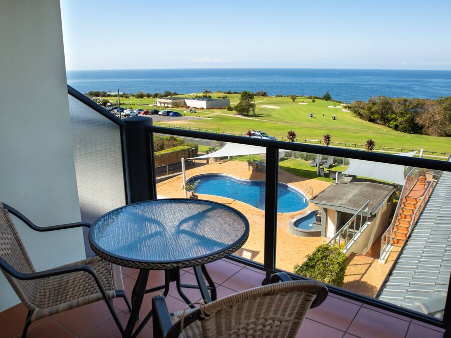 Un balcon doté de fauteuils en osier et d’une table en verre, offrant une vue sur une piscine et un terrain de golf.