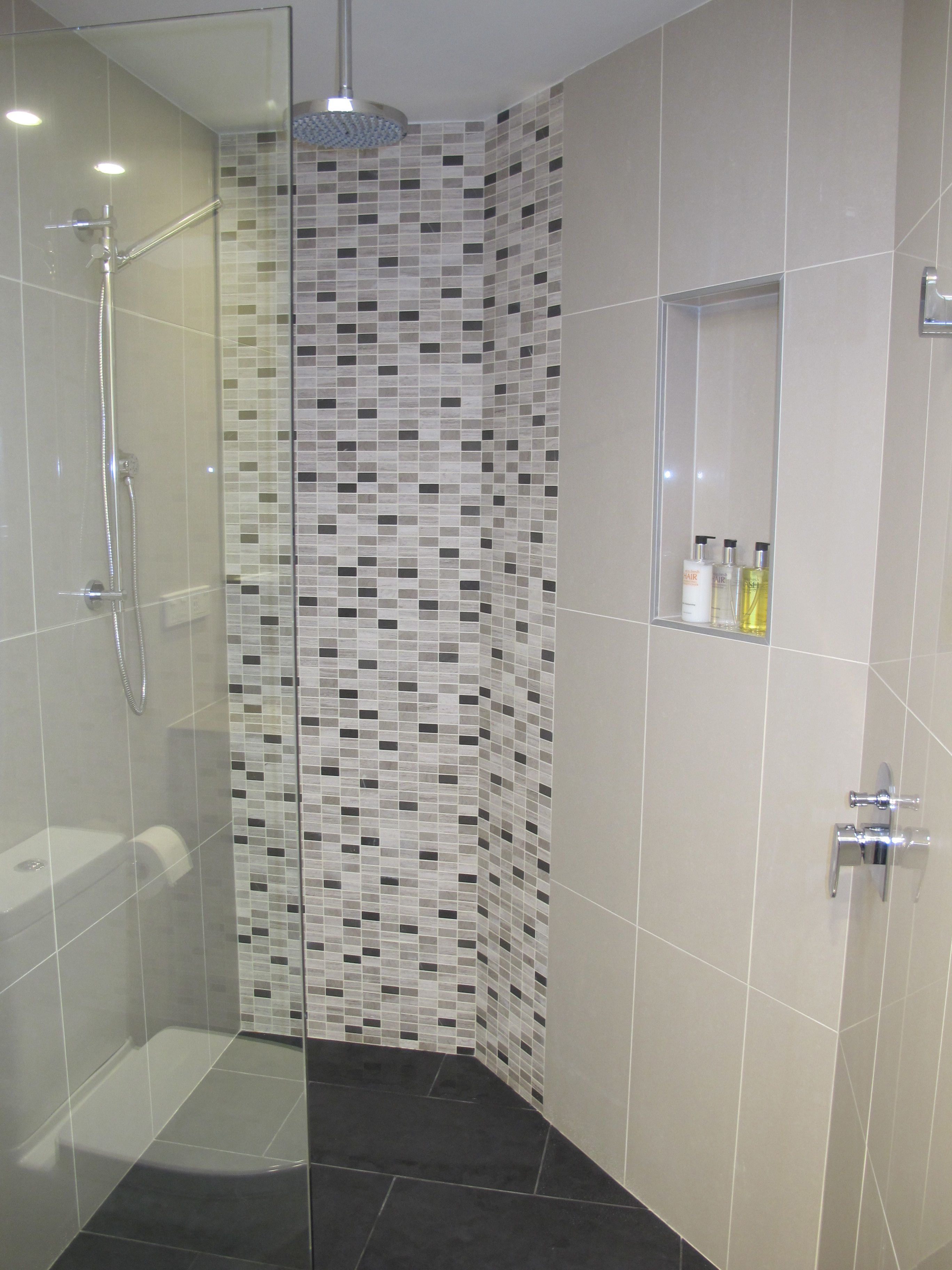 A modern shower area with a glass door, a tiled mosaic wall, and a shelf with toiletries.