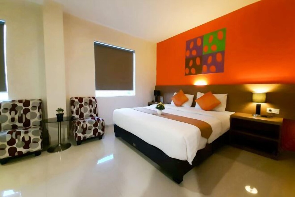 Una habitación de hotel con una cama, dos sillas, una mesita de noche, una lámpara y un ventanal.