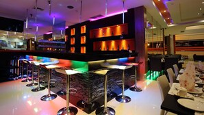 A bar area with multiple stools, a counter with shelves of bottles, and a modern ceiling design with lighting. - Miyana Hotel (Medan)