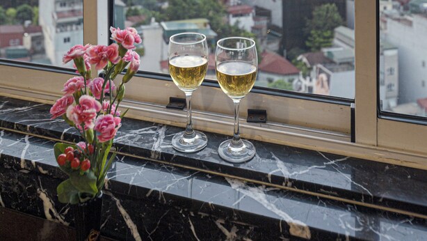 A window with a view of a cityscape, a vase with pink flowers, and two glasses of white wine on the windowsill. - Chalcedony Grand Hotel (Hanoi)