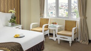 A hotel room with a bed, two striped chairs, a small table with a fruit bowl, and a view of the city through the window. - Chalcedony Grand Hotel (Hanoi)