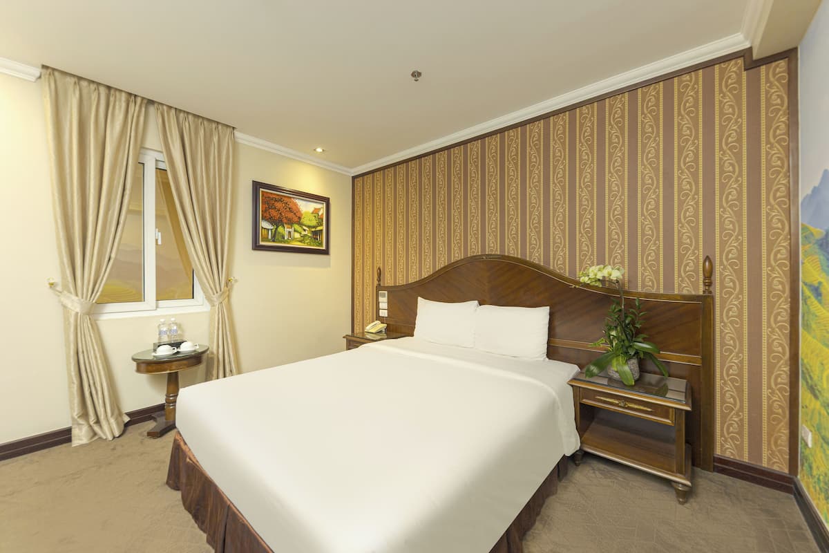 A hotel room with a large bed, a nightstand with a lamp, a small table with a vase of flowers, and a painting on the wall.