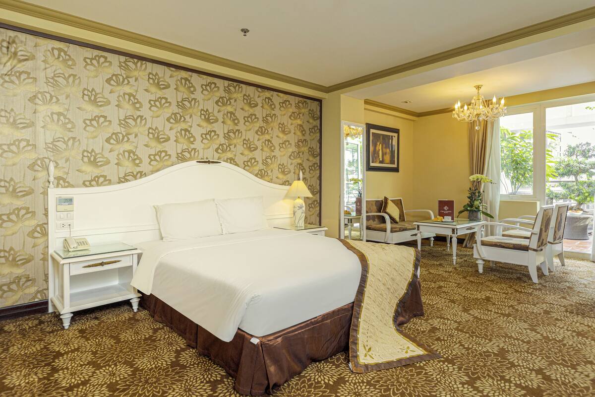 A hotel room with a large bed, a bedside table with a lamp, a chair, and a small table with a vase of flowers.