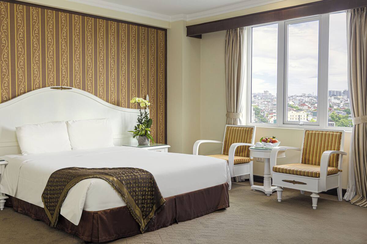 A hotel room with a large bed, two chairs, a small table, and a view of the city through the window.
