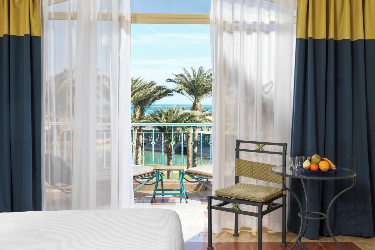 A hotel room with a bed, a chair, a small table with fruit, and a view of a pool and palm trees.
