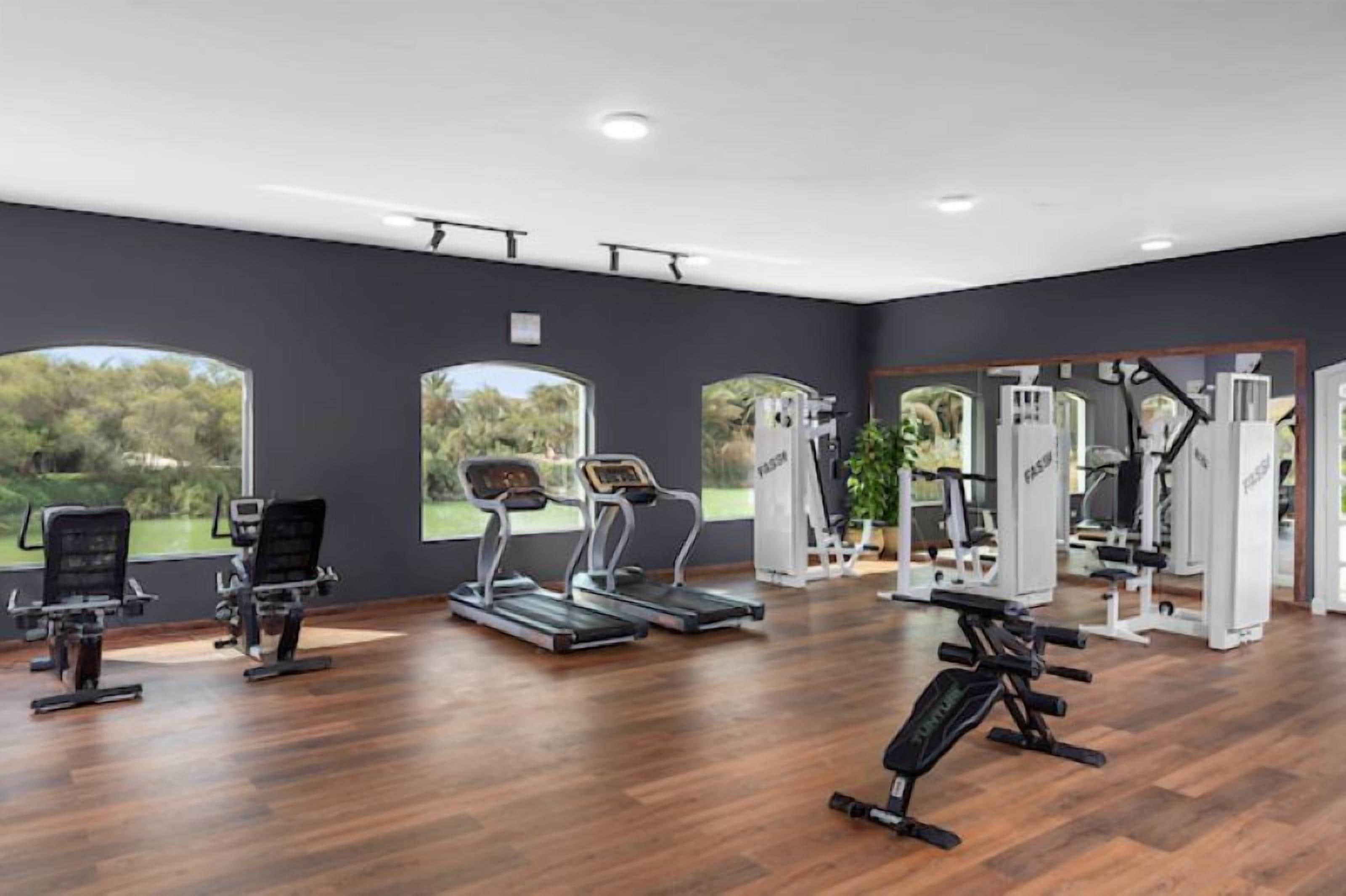 A gym with various exercise equipment, large windows, and a wooden floor.