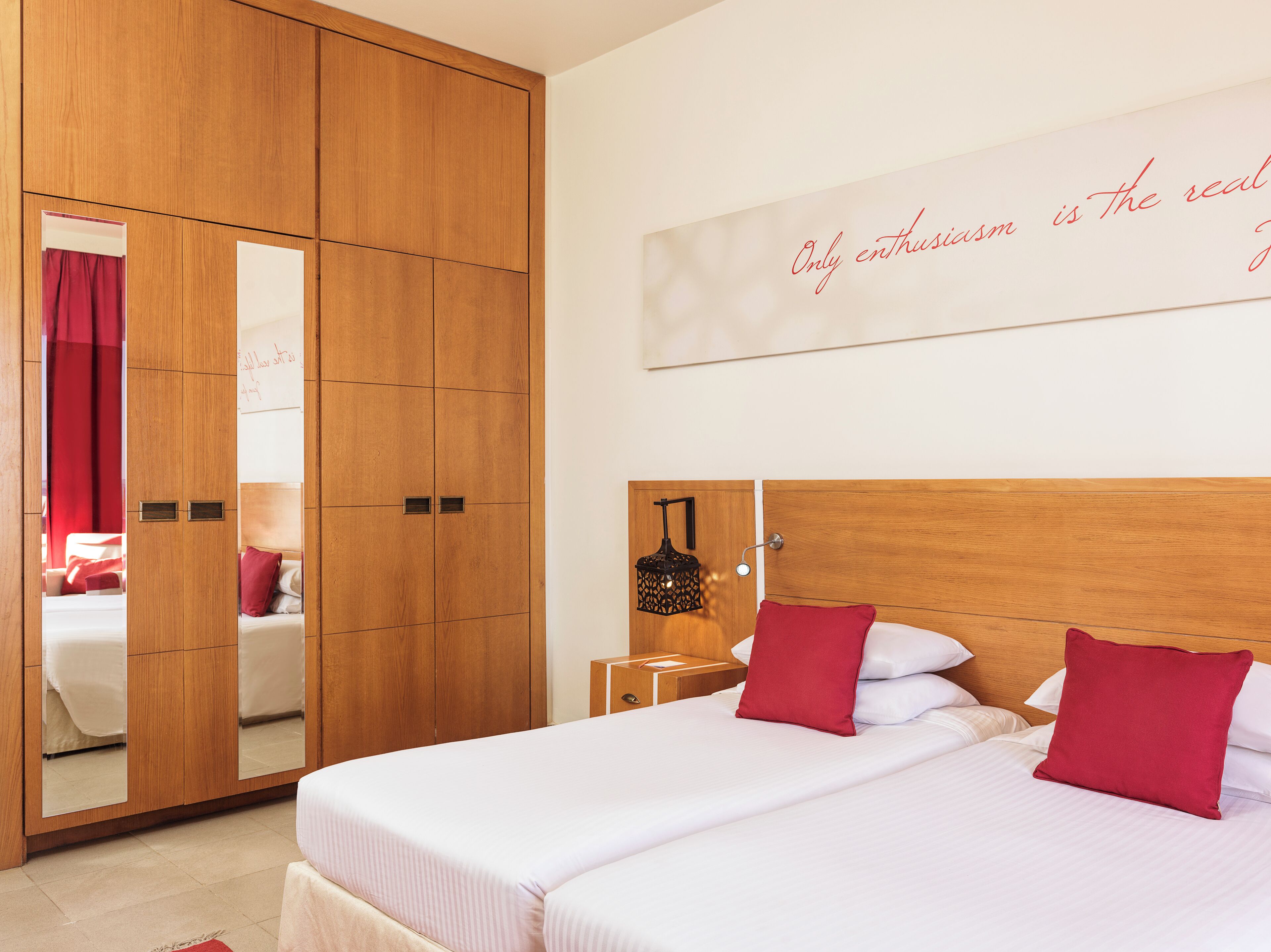 A hotel room with a large wardrobe, a bed with red pillows, a nightstand with a lamp, and a wall with a motivational quote.
