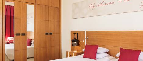 A hotel room with a large wardrobe, a bed with red pillows, a nightstand with a lamp, and a wall with a motivational quote.