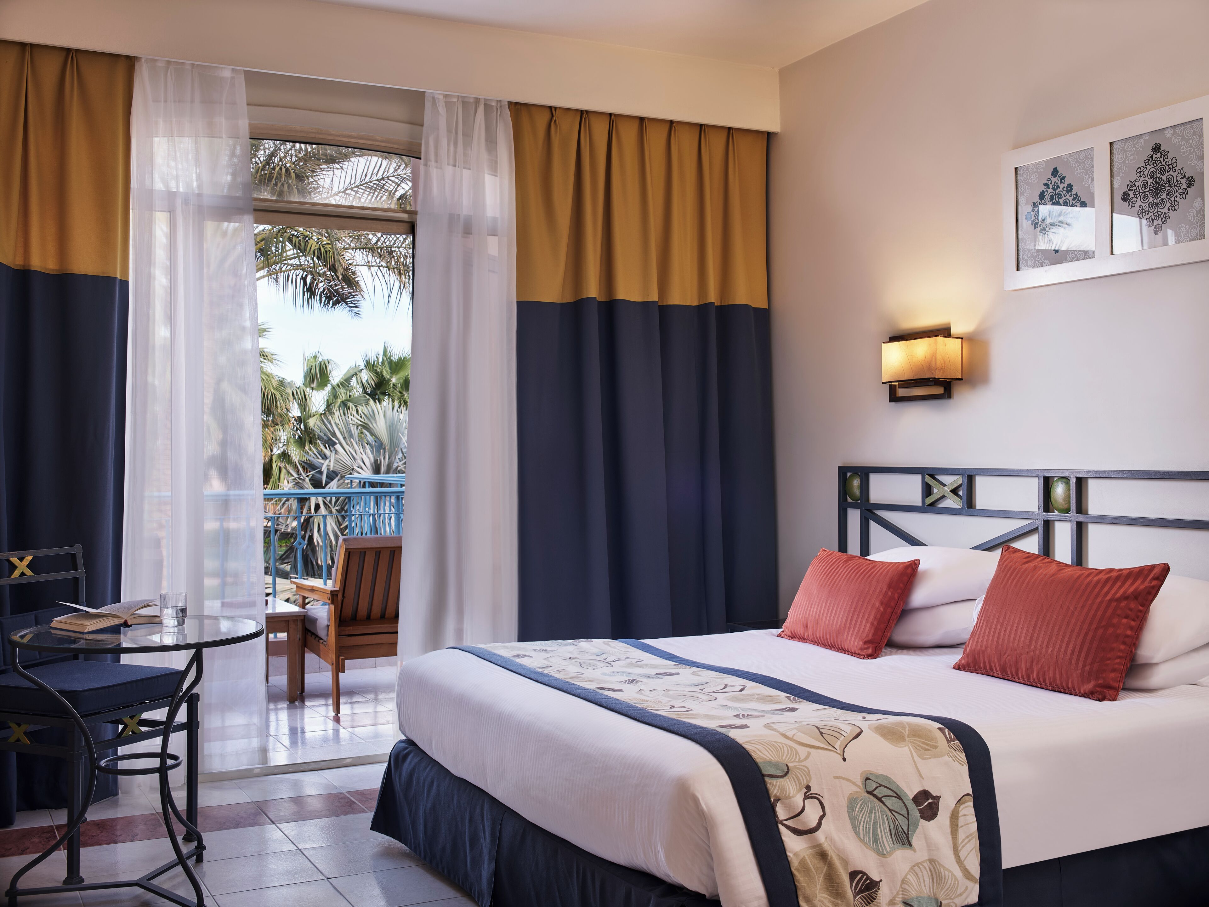 A hotel room with a bed, a desk with a chair, a balcony with a view of palm trees, and a wall-mounted light fixture.