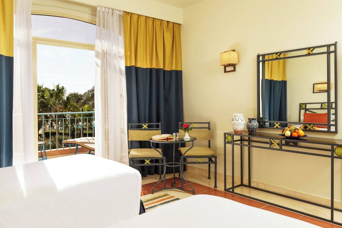 A hotel room with a bed, a desk with a mirror, a chair, a small table with a vase and fruit, and a balcony with a railing.
