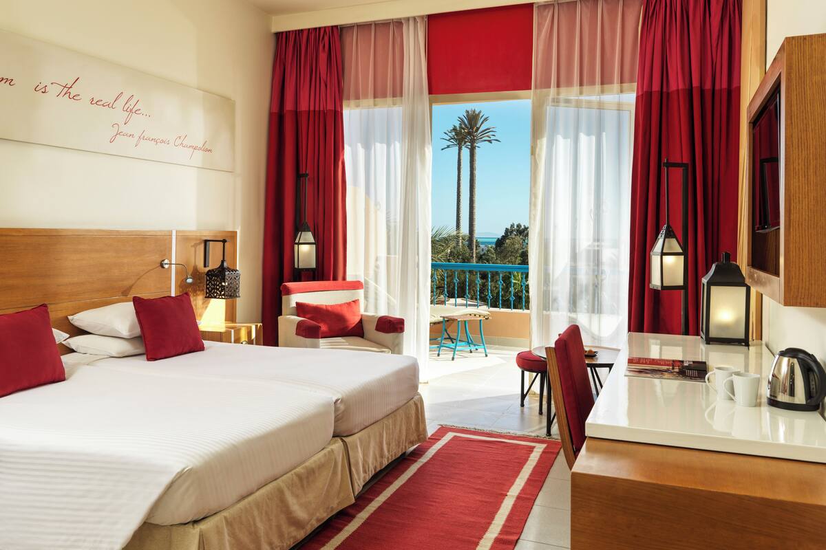 A hotel room with a large bed, a desk with a chair, a balcony with a view, and a red and white color scheme.